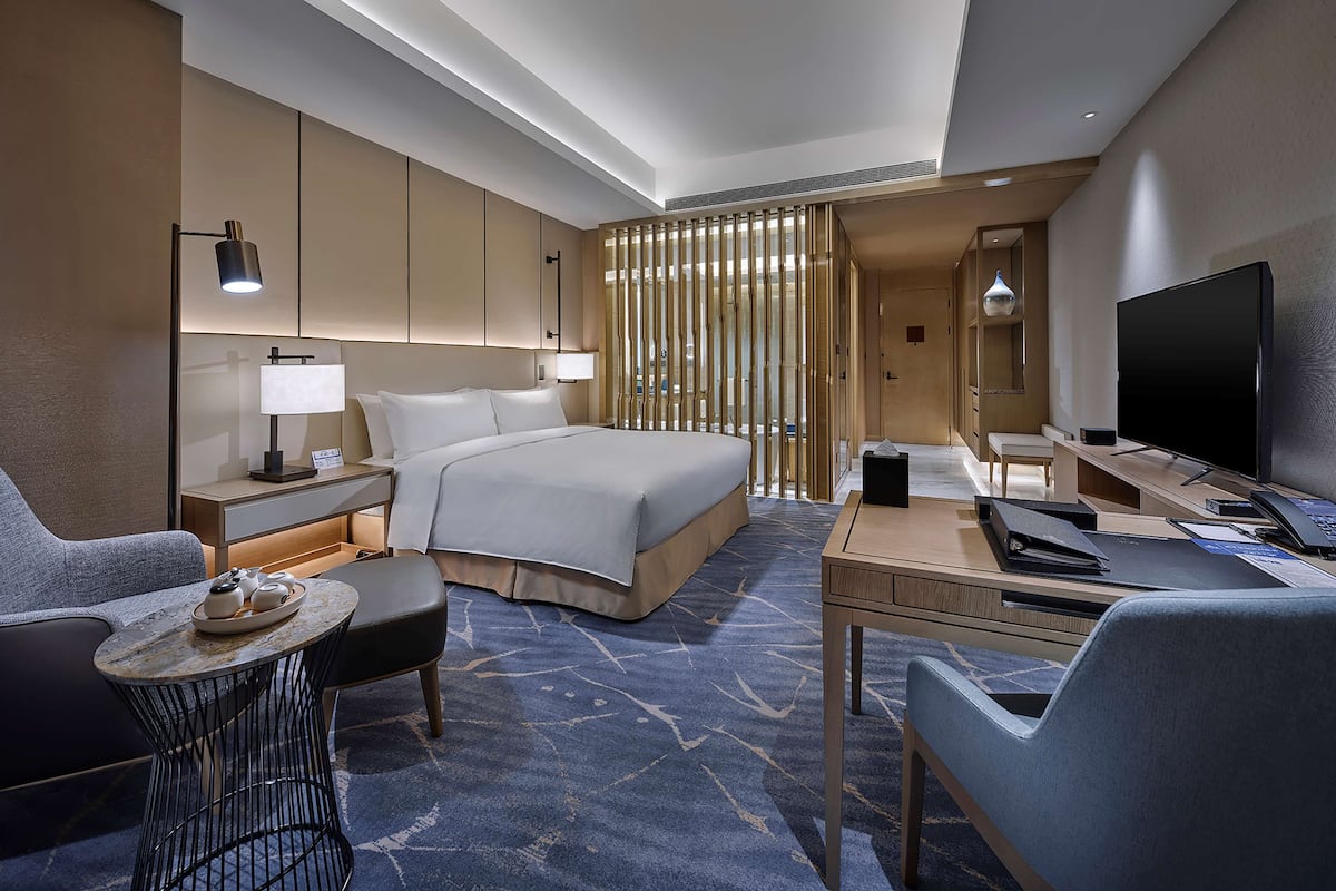 Una habitación de hotel moderna con una cama grande, un escritorio con televisor de pantalla plana, una zona de estar con una silla y una mesita, y una separadora decorativa.