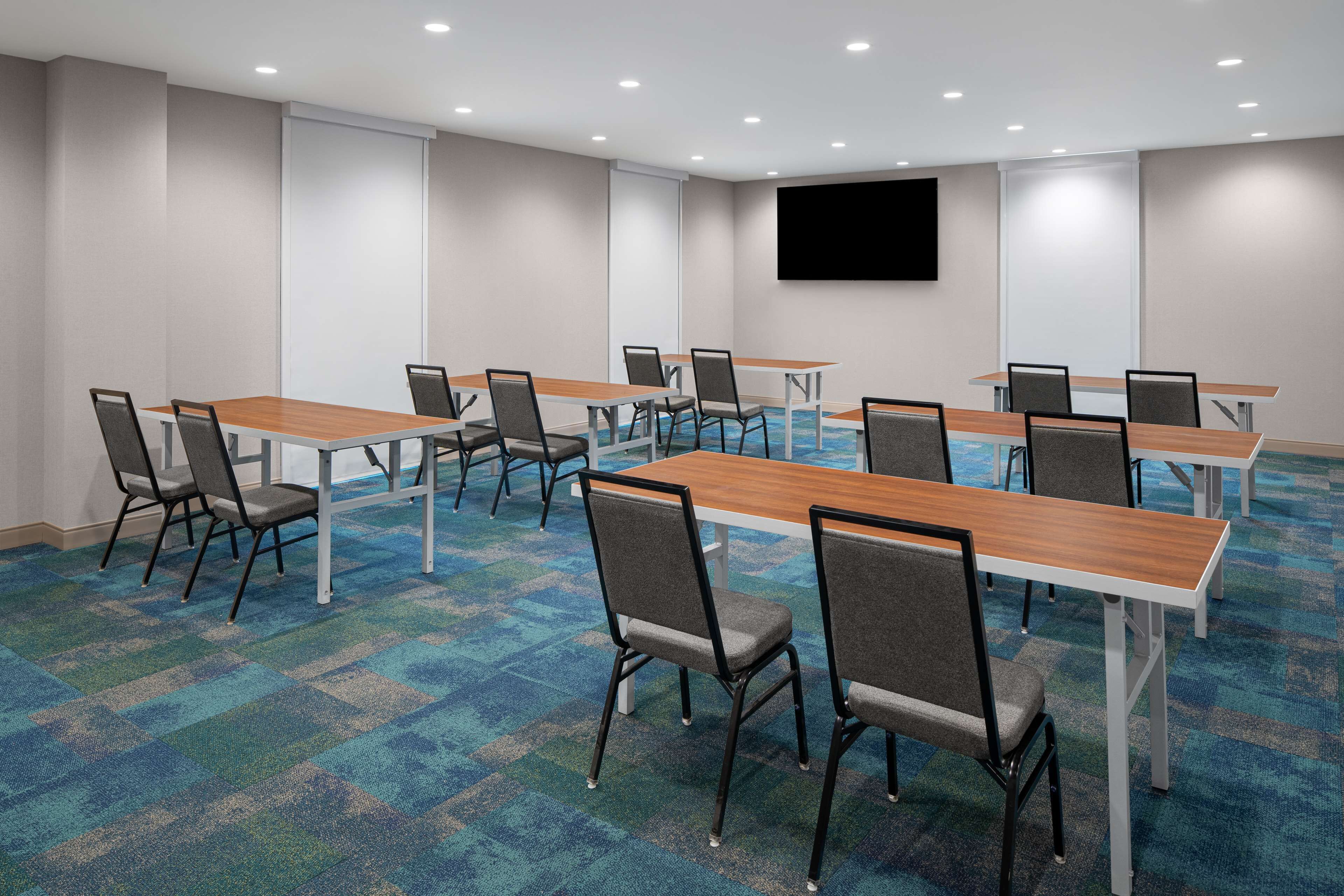 A conference room with multiple tables and chairs, a flat-screen TV mounted on the wall, and a carpeted floor.