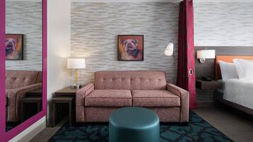A hotel room with a sofa, a round ottoman, a bed, a nightstand, a lamp, a wall-mounted TV, and a mirror.