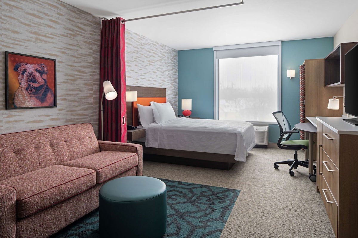 Ein Hotelzimmer mit Bett, Sofa, Schreibtisch mit Stuhl, Fernseher und einem Fenster mit Vorhängen.