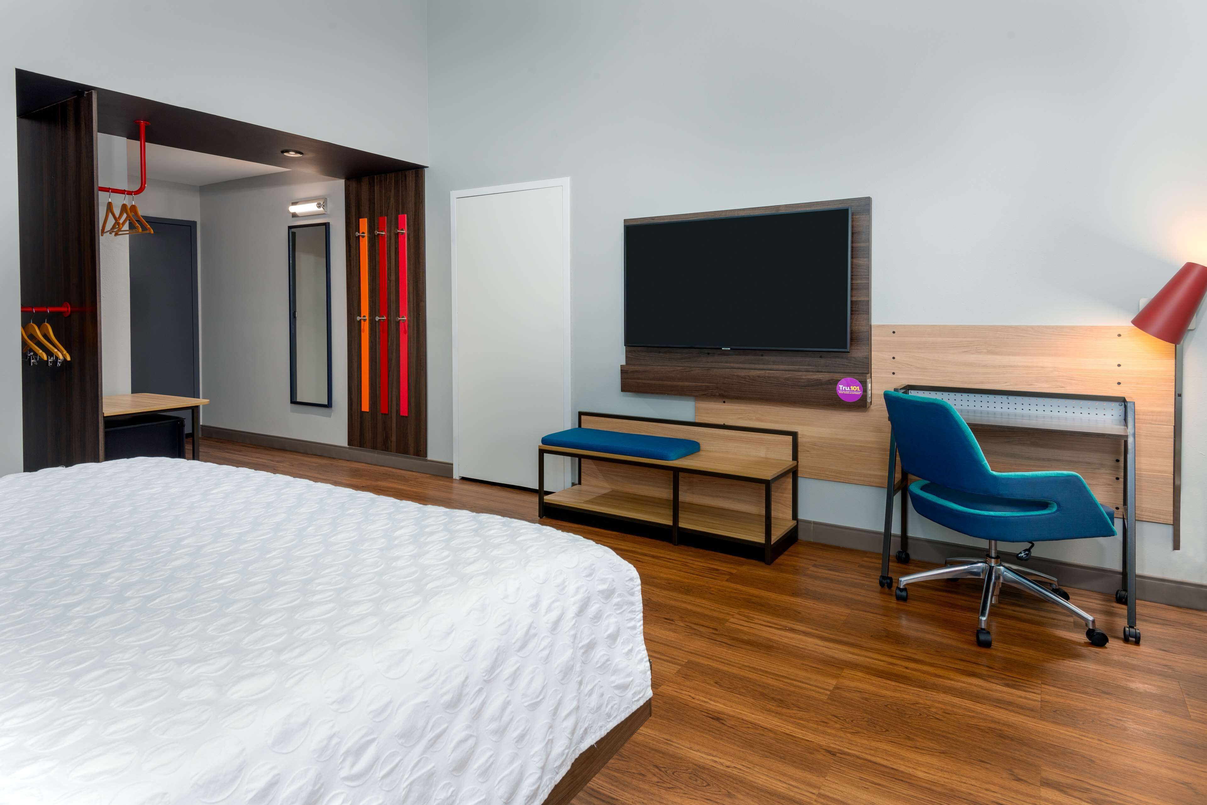 A modern hotel room with a large bed, a desk with a chair, a flat-screen TV, and a red lamp.