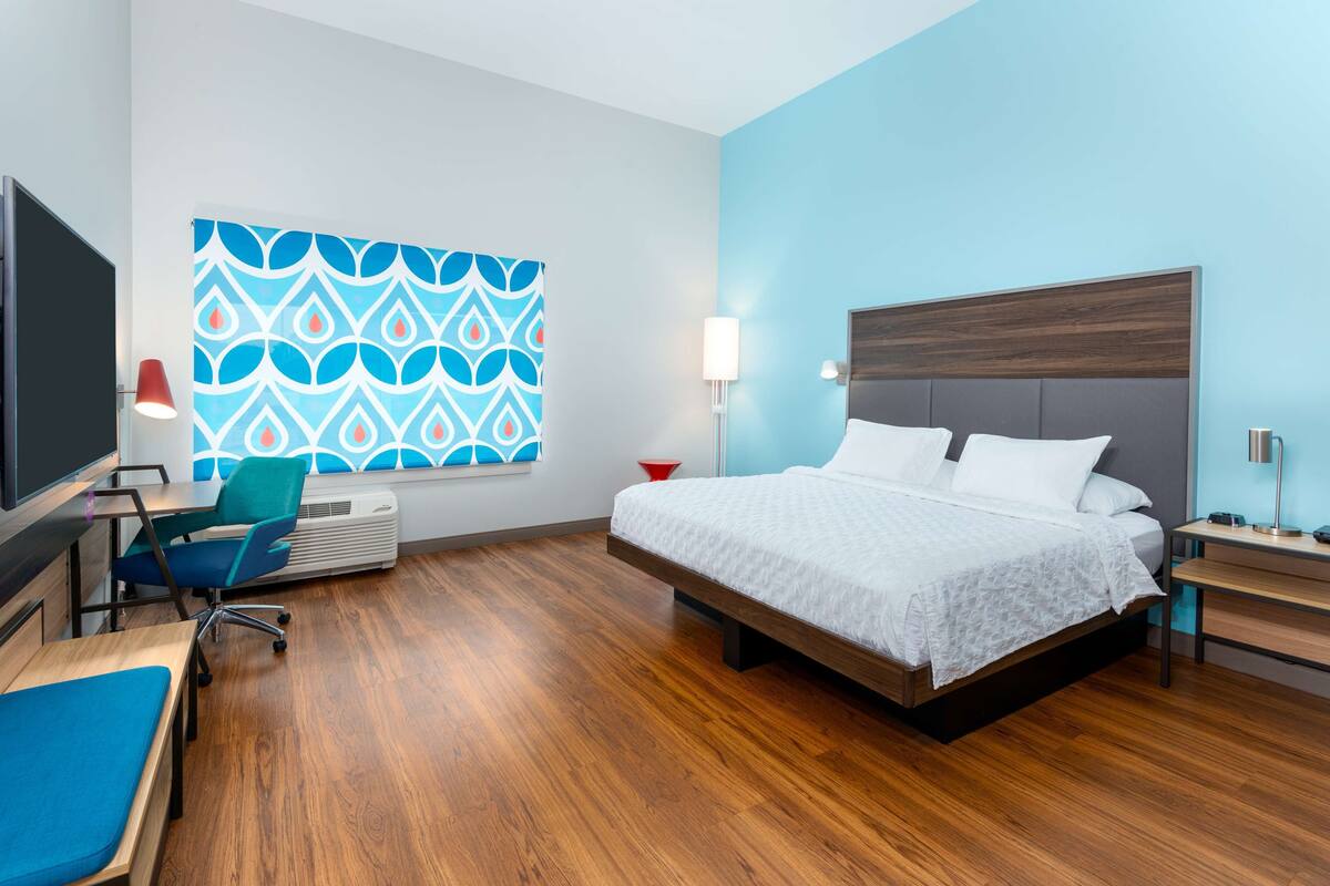 Ein modernes Hotelzimmer mit Holzboden, einem großen Bett mit weißer Bettwäsche, einem Schreibtisch mit Stuhl, einem Flachbildfernseher und einer Wand-Klimaanlage.