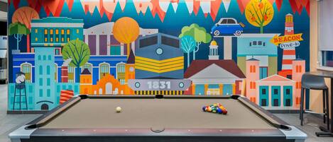 Una mesa de billar con un mural de colores al fondo que muestra un paisaje urbano y la palabra "HURRICUT".