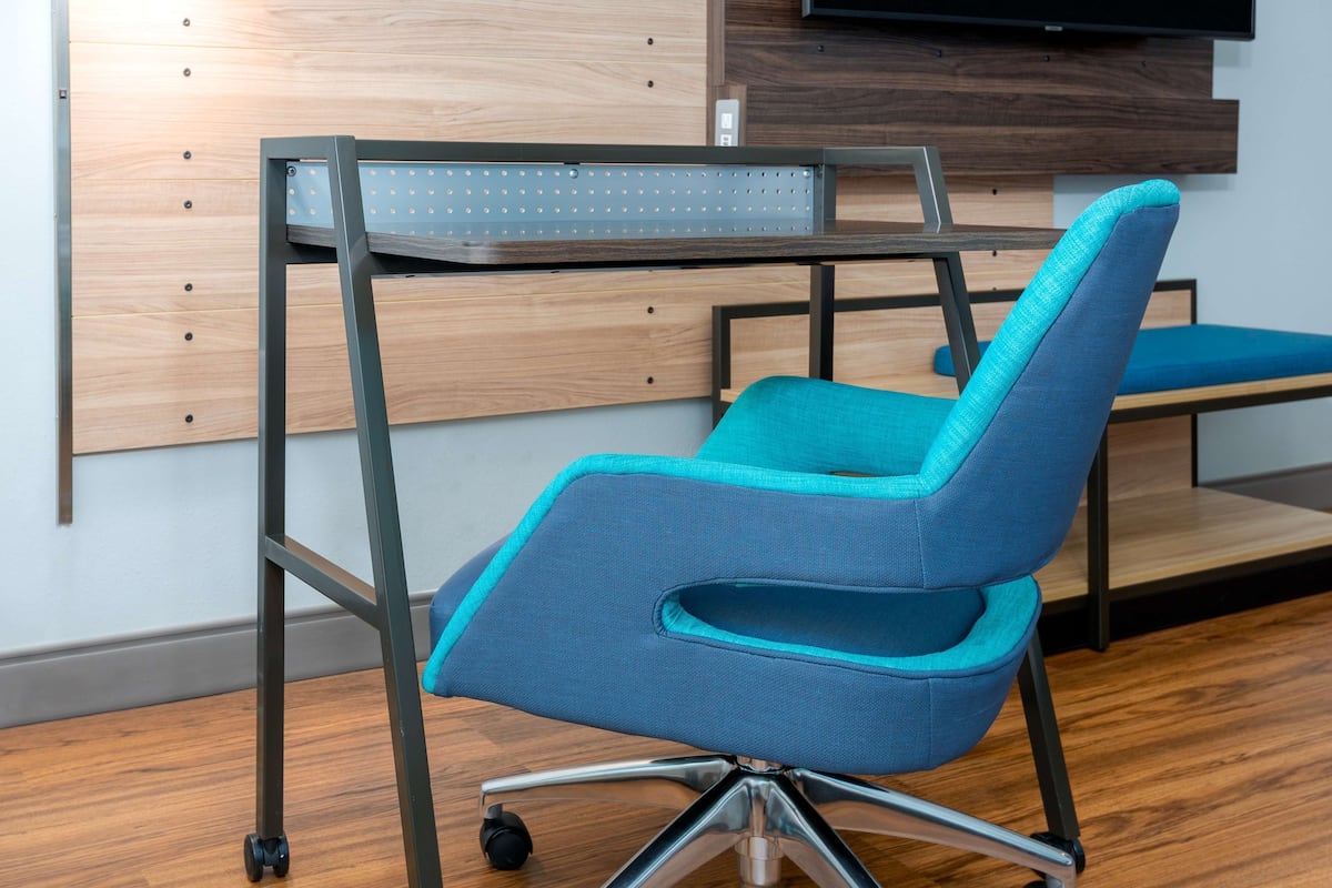Ein moderner Bürostuhl mit blauer Polsterung und Metallgestell, vor einer Holzwand mit einem Nagelbrett und einem angebrachten Fernseher.