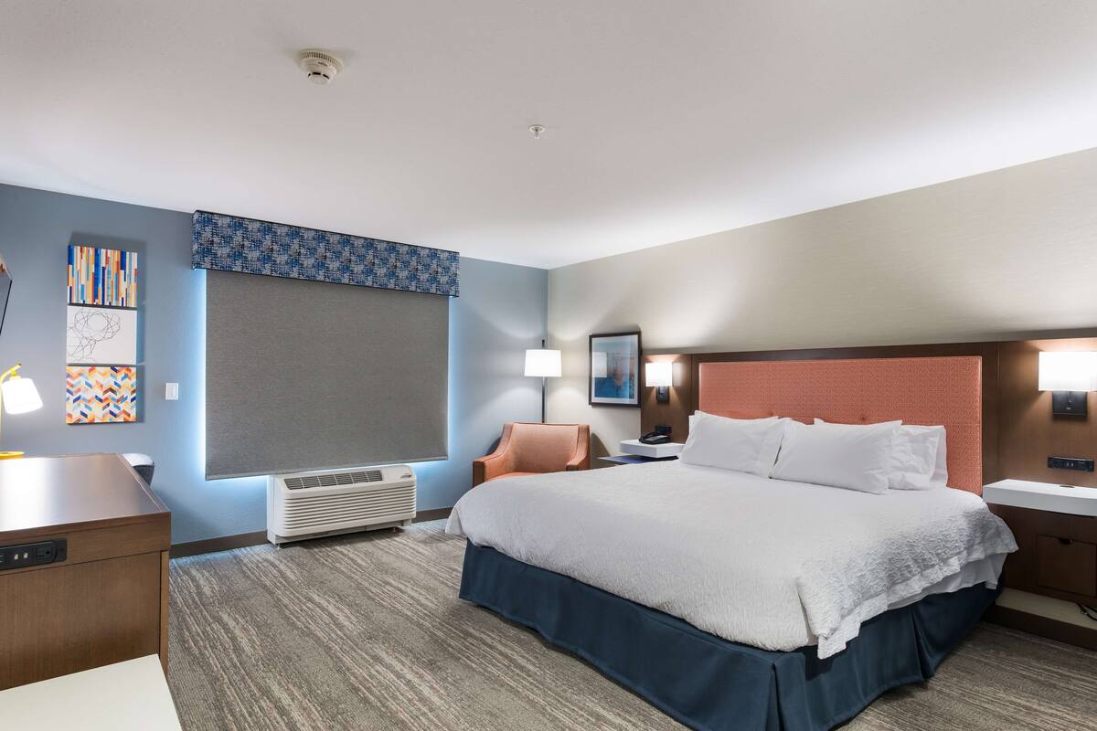 A hotel room with a large bed, a desk, a chair, a nightstand, a lamp, and a wall-mounted air conditioner.