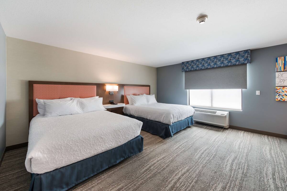 A hotel room with two beds, a large window with a view, a ceiling-mounted air conditioner, and a wall-mounted artwork.