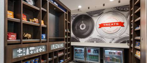 A snack and beverage area with shelves stocked with various snacks and drinks, a clock, and a wall mural with a 'Treats' sign.