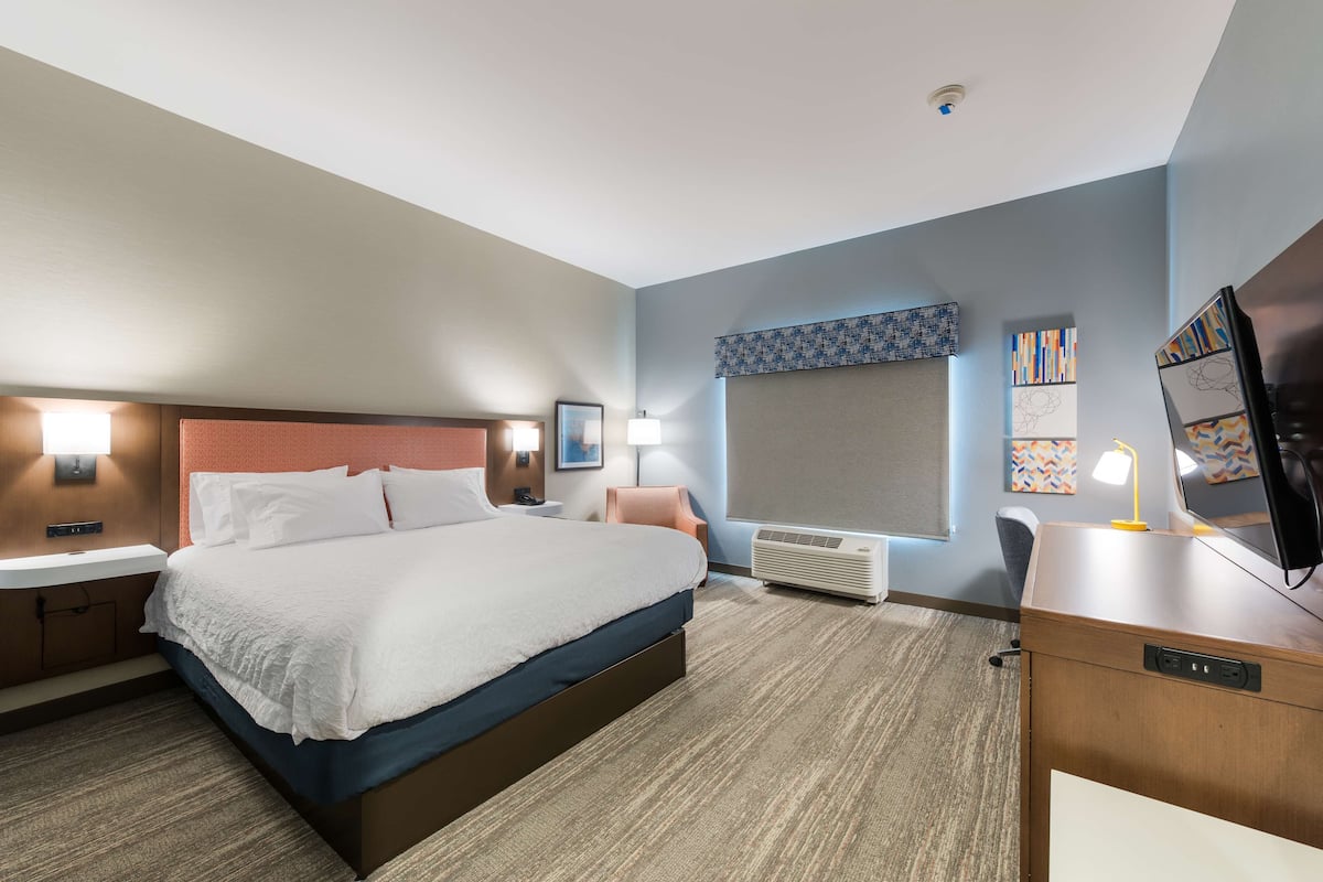 A hotel room with a large bed, bedside tables, a desk with a computer, a window with blinds, and artwork on the walls.