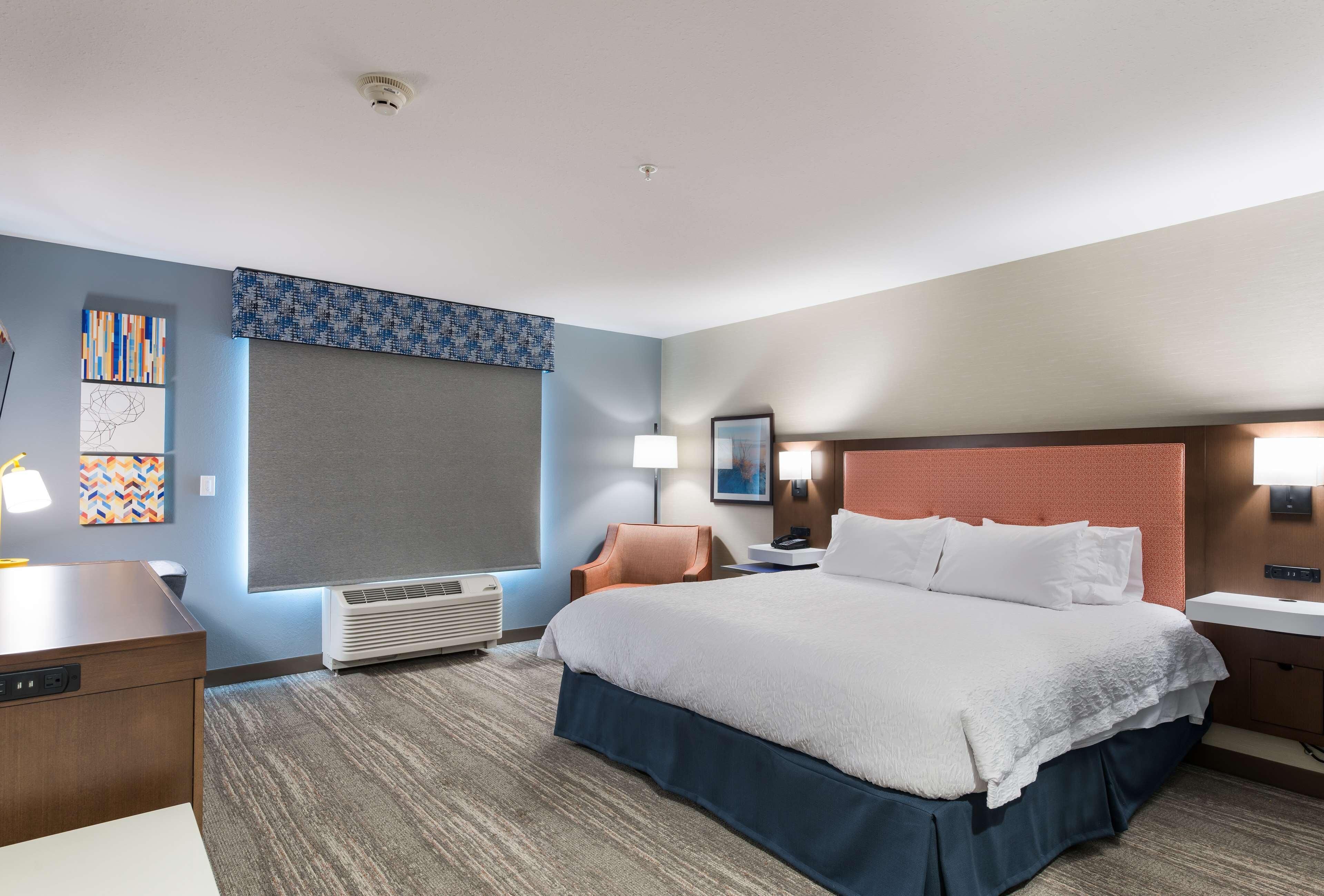 A hotel room with a bed, a desk, a chair, a lamp, a nightstand, and a wall-mounted air conditioner.