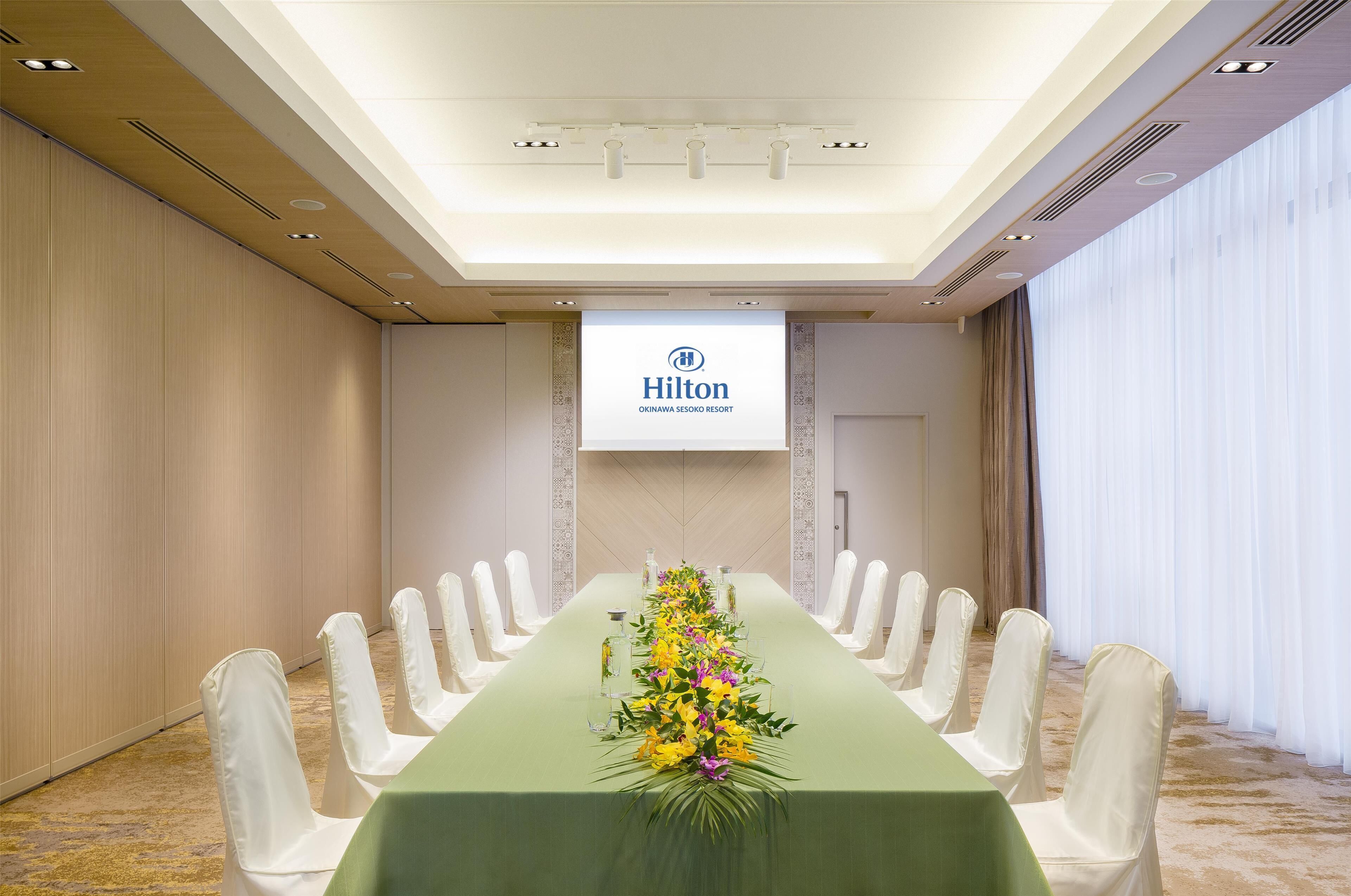Una sala de conferencias con una mesa larga, sillas blancas y un centro de mesa floral. Se ve el logotipo de Hilton en la pared.