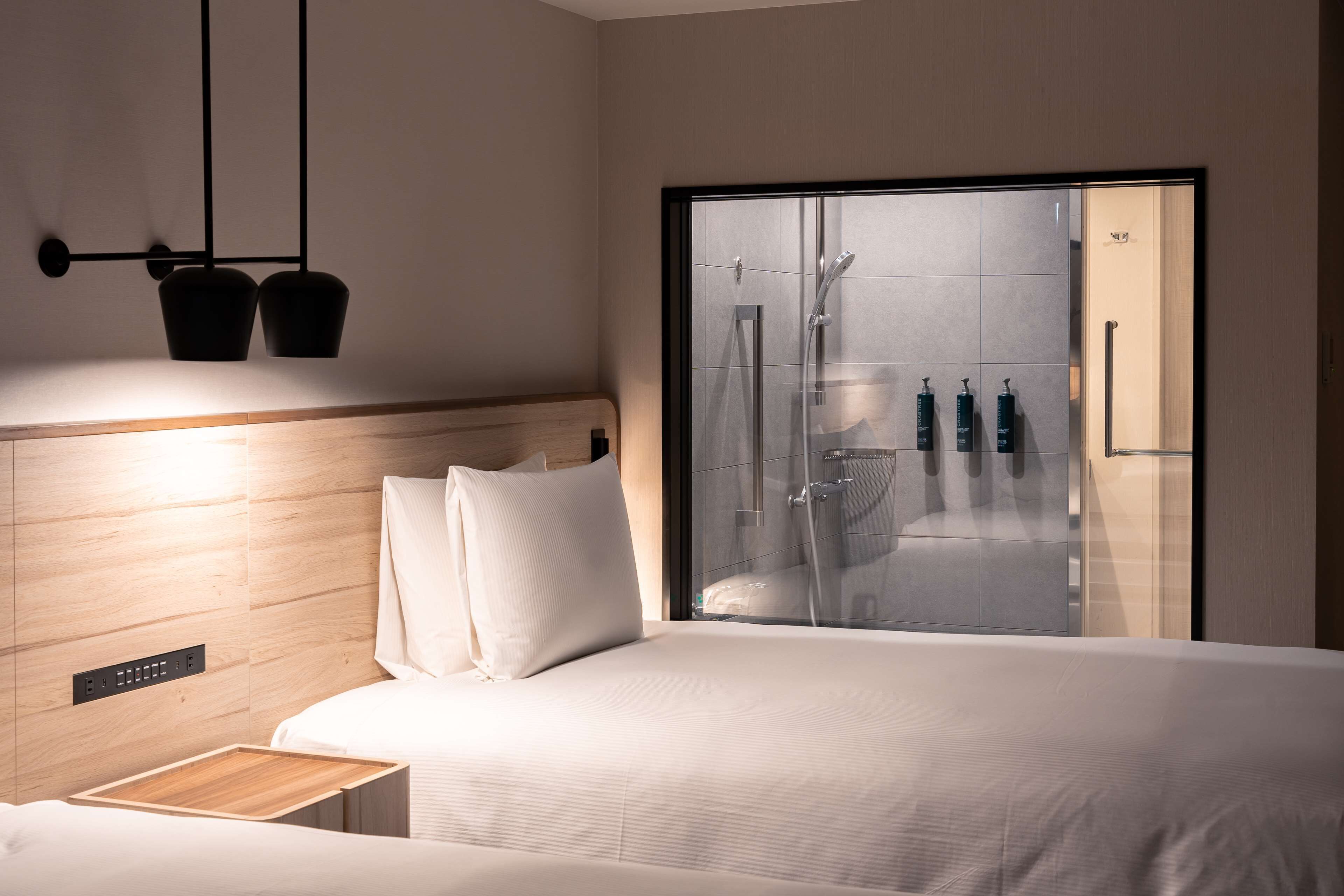 A modern hotel room with a wooden headboard, a large bed, a bedside table, and a glass shower enclosure with toiletries.