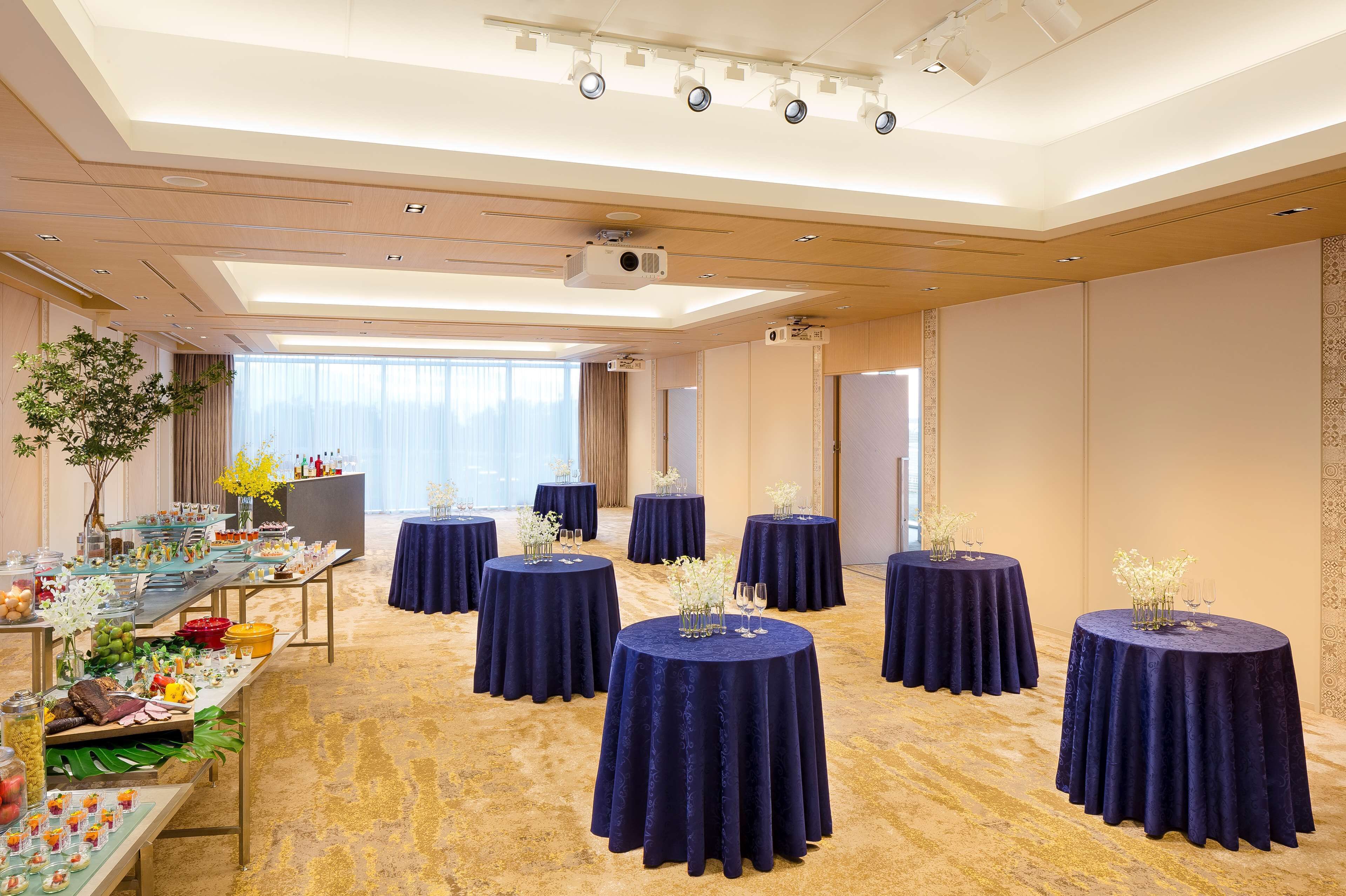 Un salón de conferencias con mesas redondas cubiertas con manteles azul oscuro, una zona de buffet con diversas opciones de comida y un ventanal que proporciona luz natural.