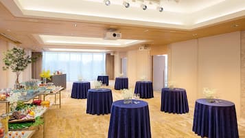 Un salón de conferencias con mesas redondas cubiertas con manteles azul oscuro, una zona de buffet con diversas opciones de comida y un ventanal que proporciona luz natural.