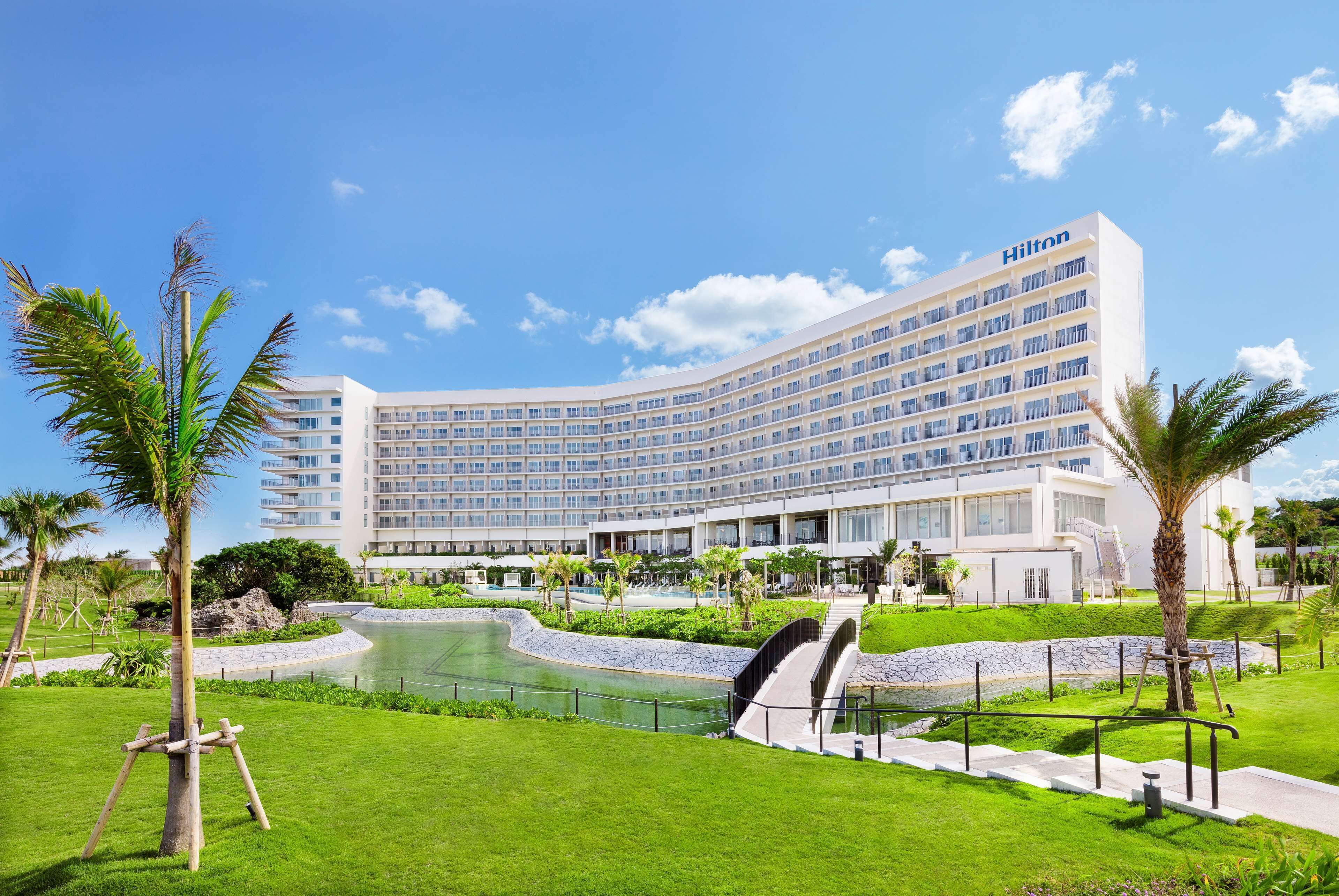 A large, modern hotel with a curved facade, a swimming pool, and well-maintained green lawns.