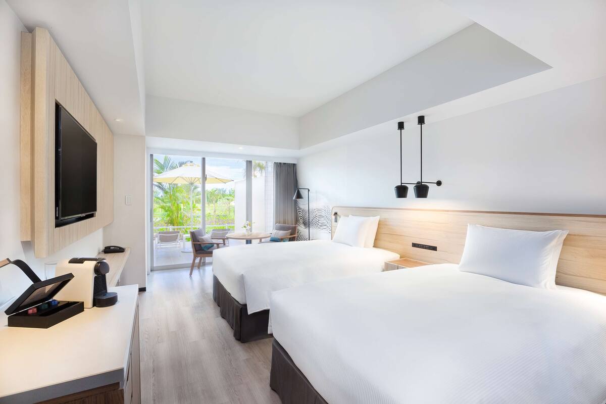 Habitación de hotel con dos camas, televisión, escritorio con lámpara y vista al exterior.