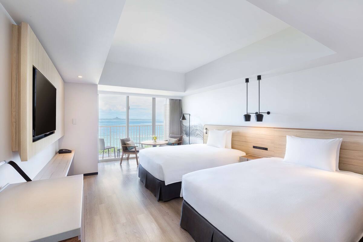 Habitación de hotel con dos camas, televisión, escritorio y balcón con vista al mar.