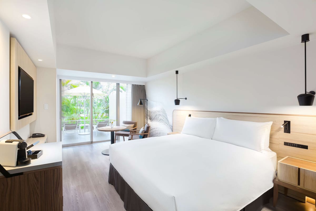 Una habitación de hotel moderna con una cama grande, un escritorio con computadora, televisor y un balcón con mobiliario exterior.