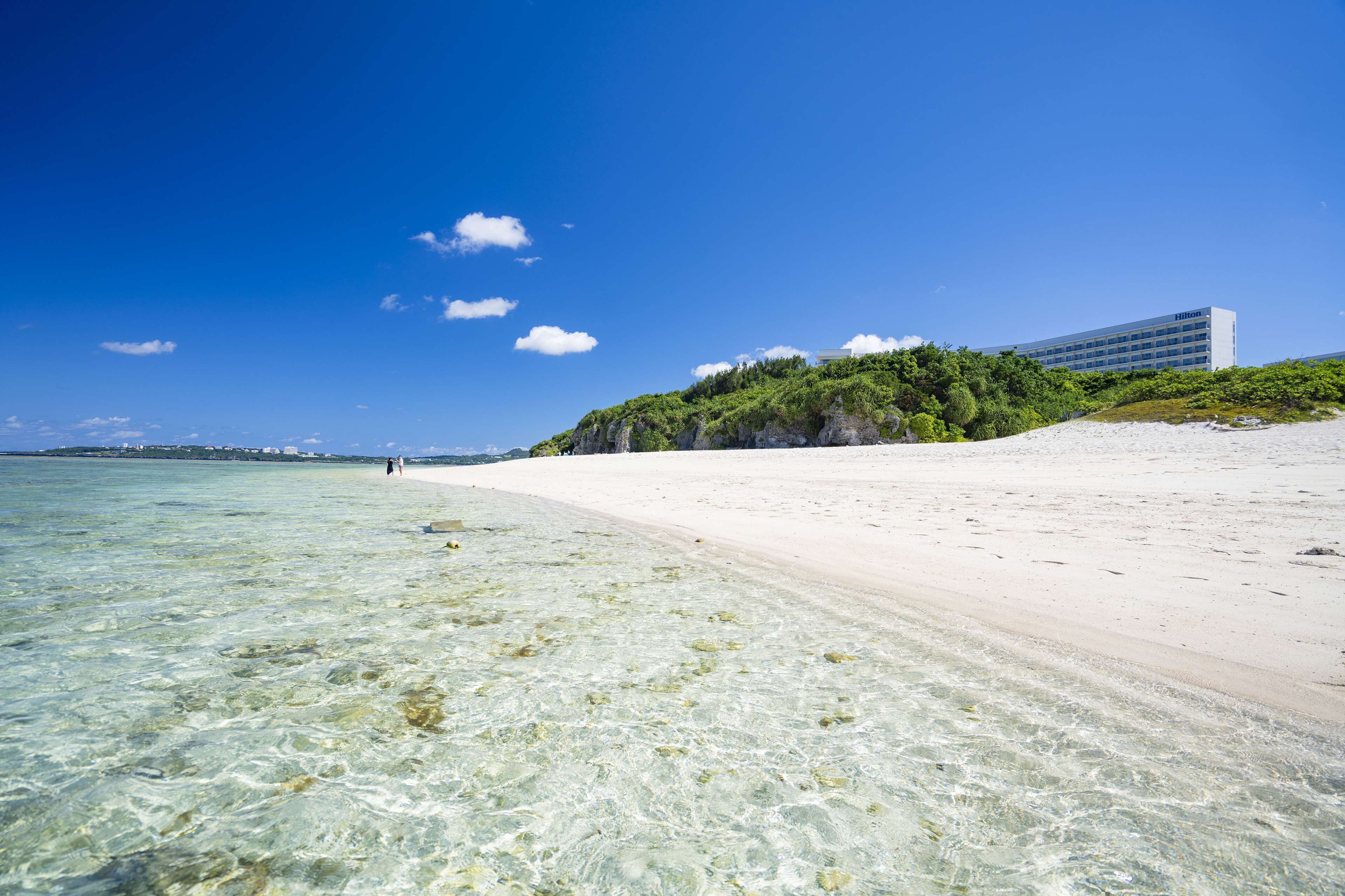 A beach with clear shallow waters, a sandy shore, and a building with a flat roof in the background.