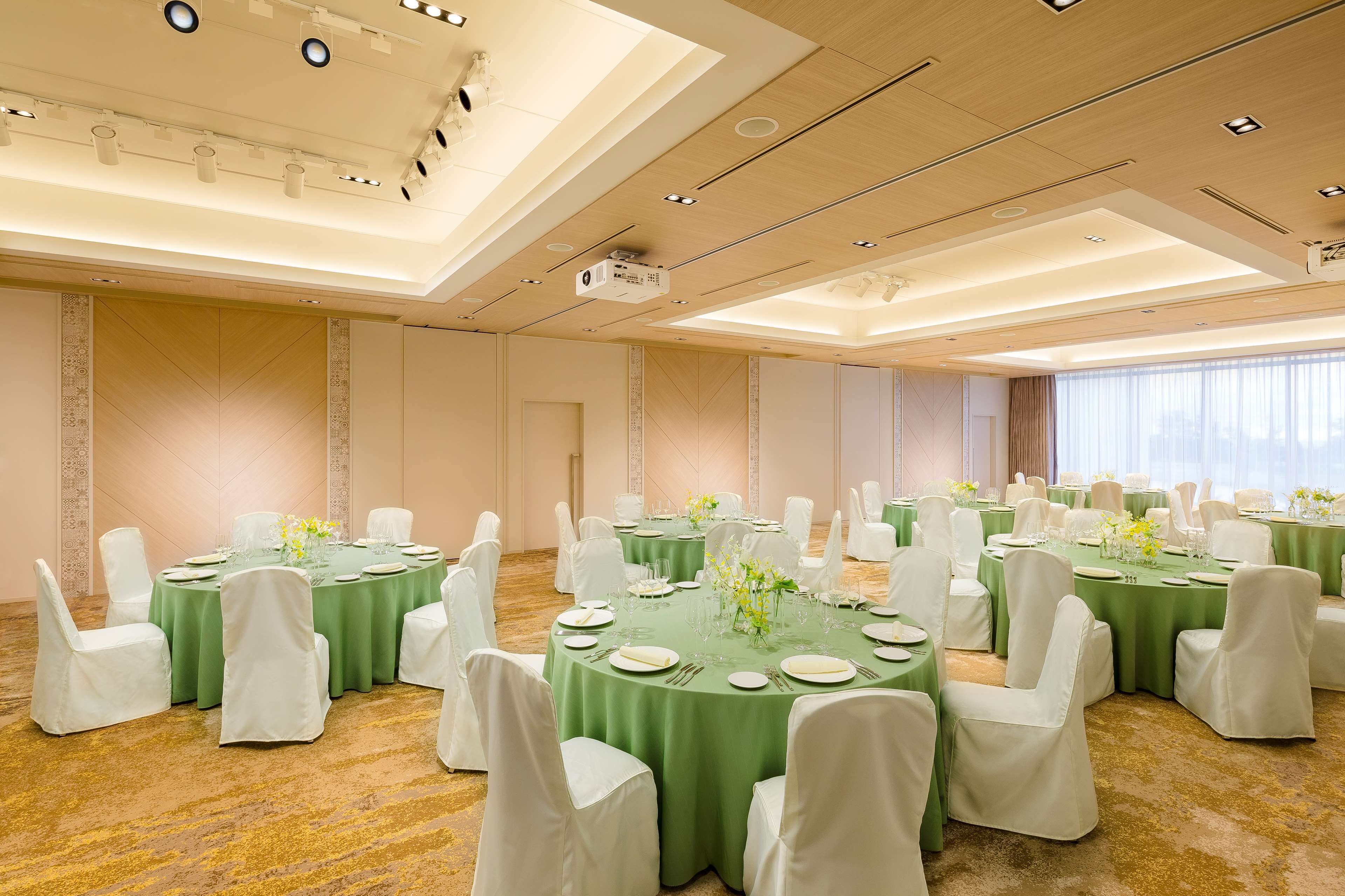 Un salón de conferencias con varias mesas redondas, cada una con sillas blancas y manteles verdes, decorado con centros de mesa florales y vajilla.