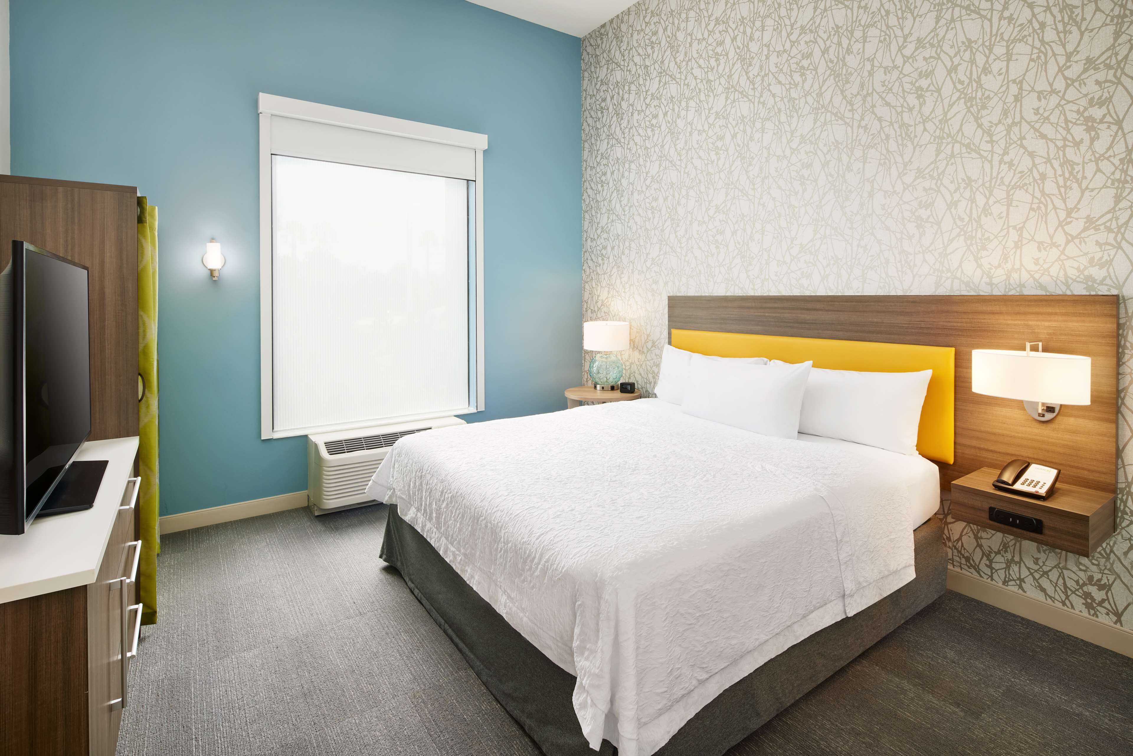 A hotel room with a bed, a desk with a TV, a window, and a wall with a patterned design.