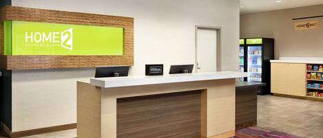 A hotel reception desk with a 'HOME 2' sign, a minibar, and a 'Cold' refrigerator.