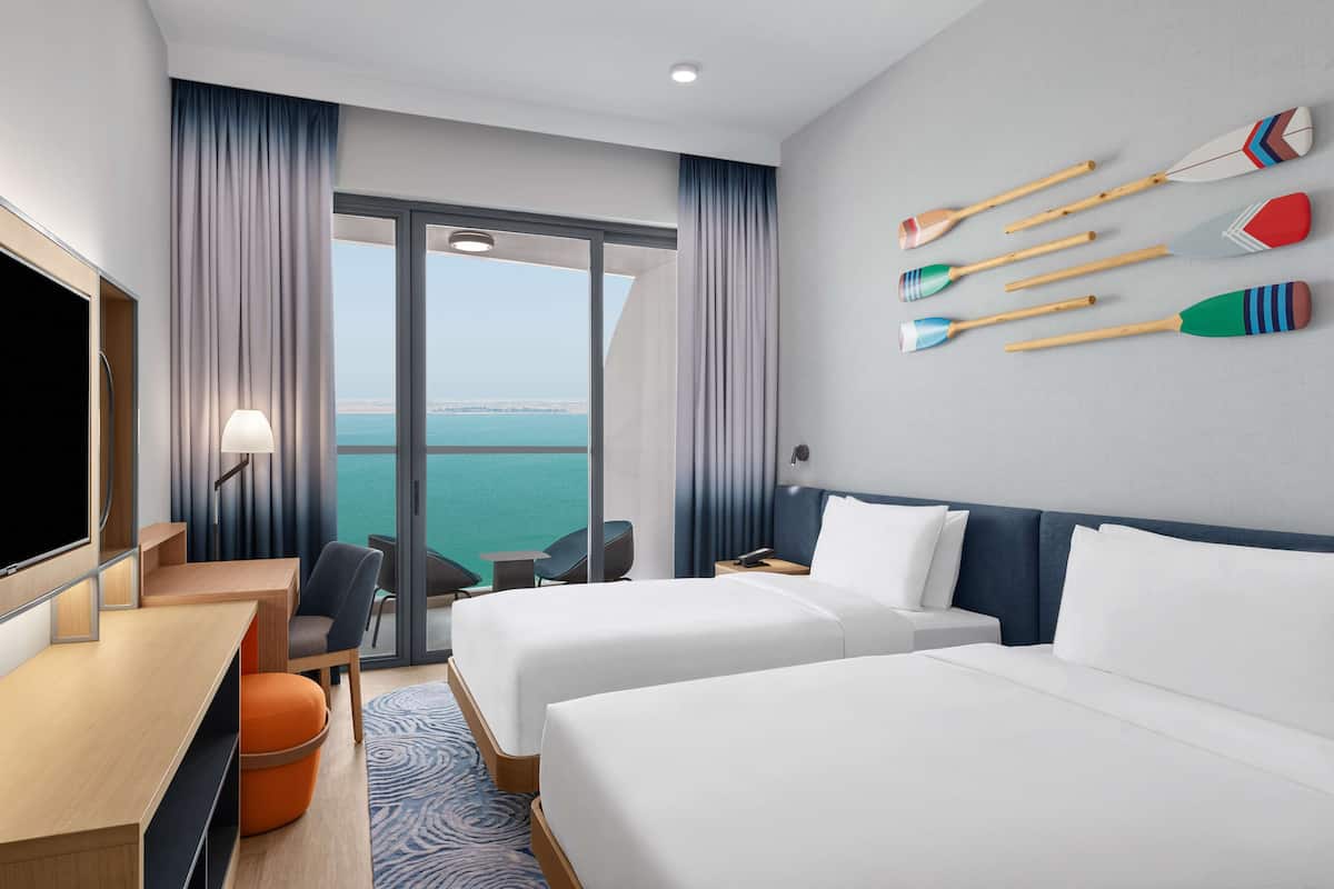 Ein Hotelzimmer mit zwei Betten, einem Schreibtisch, einem Stuhl, einem Fernseher und Blick aufs Meer.