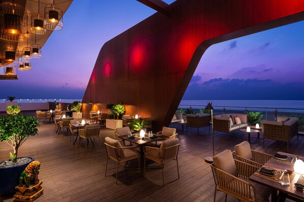 An outdoor dining area with wooden tables and chairs, potted plants, and a view of the ocean at dusk.