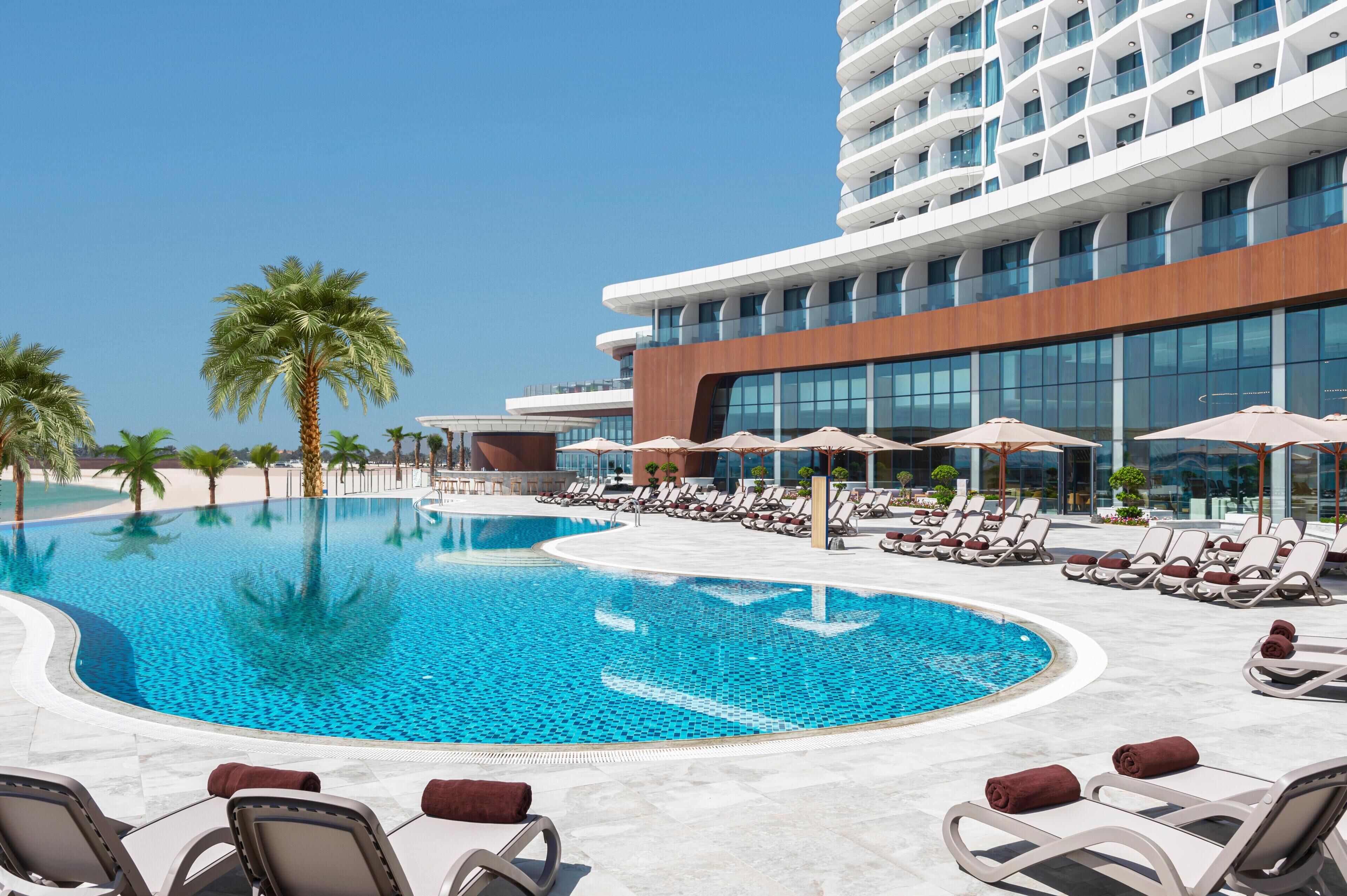 A modern hotel with a swimming pool, sun loungers, and palm trees.