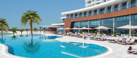 A modern hotel with a swimming pool, sun loungers, and palm trees.