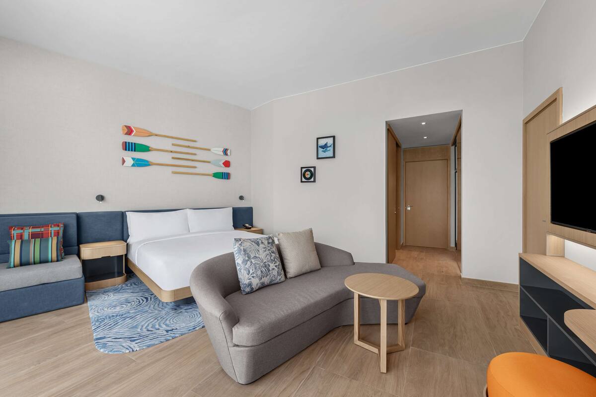 Ein modernes Hotelzimmer mit einem Bett, einem grauen Sofa, einem kleinen runden Tisch und einem Fernseher.