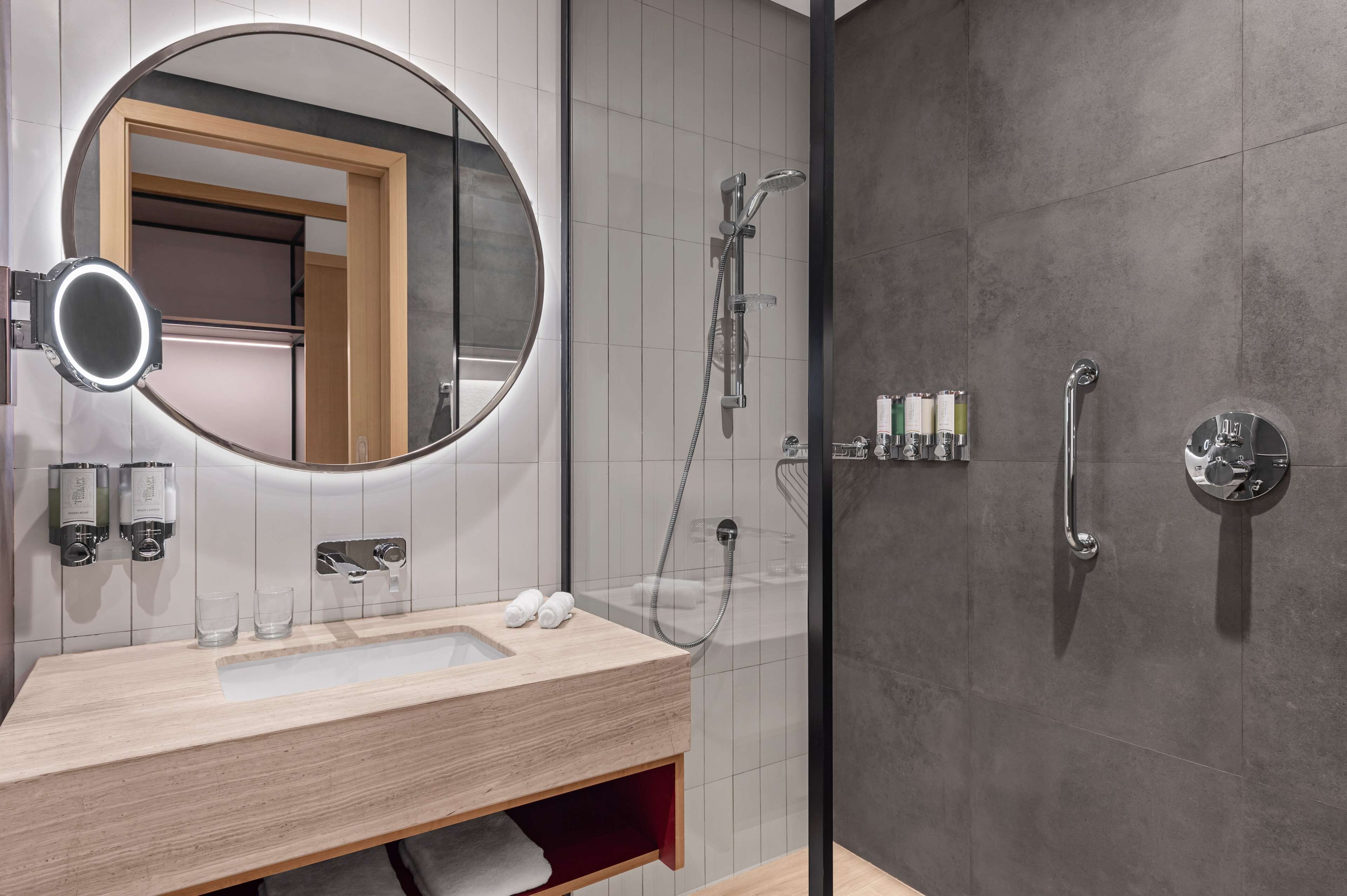A modern bathroom with a round mirror, a sink with two glasses, a handheld showerhead, and a glass-enclosed shower.