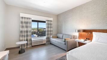 Een moderne hotelkamer met een groot bed, een slaapbank, een klein tafeltje en een raam met gordijnen.