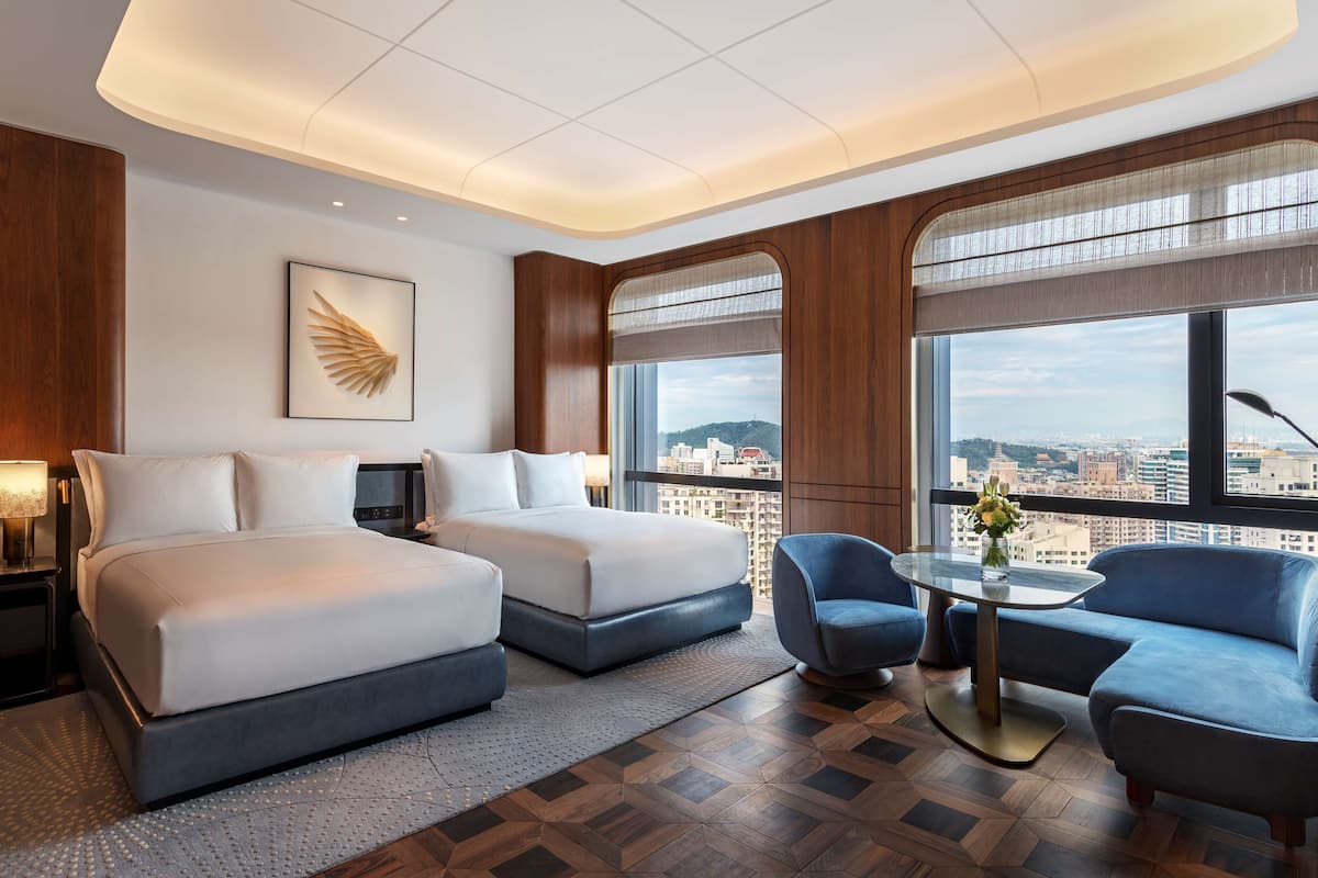 Ein Hotelzimmer mit zwei Betten, einer Sitzecke mit Tisch und Stühlen, großen Fenstern mit Blick auf die Stadt und einer Wanddekoration.