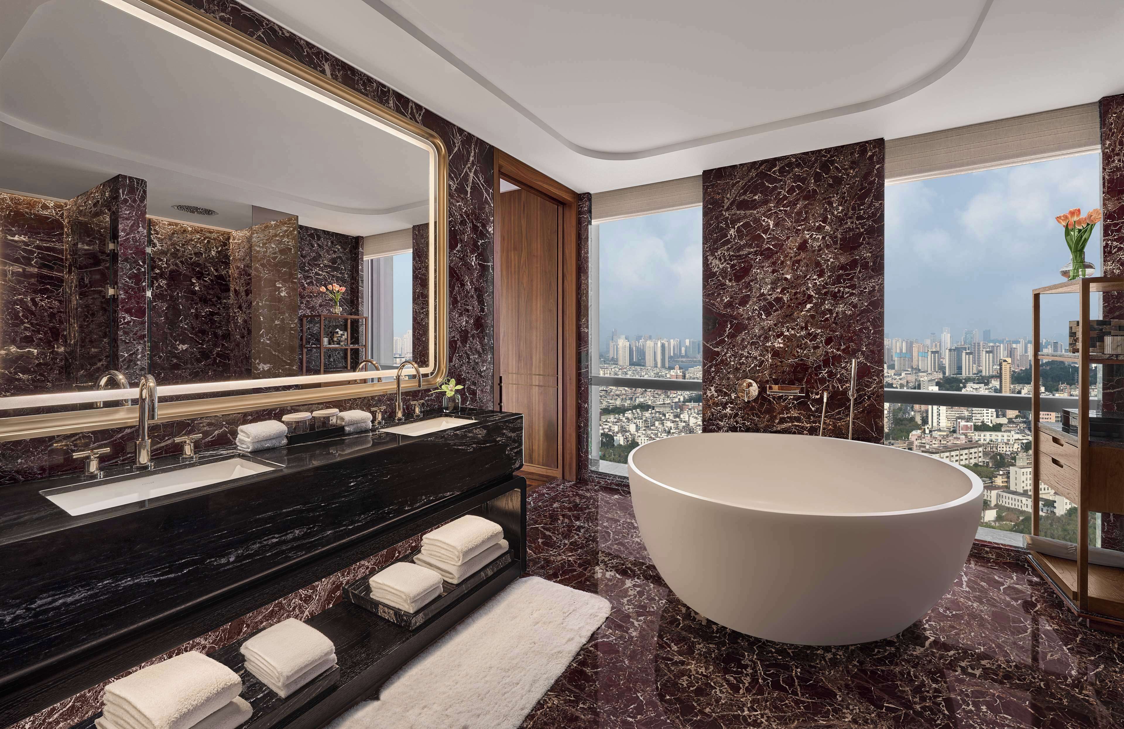 A modern bathroom with a large bathtub, a double sink vanity, and a large mirror. The room features dark marble walls and floors, and a city view through large windows.
