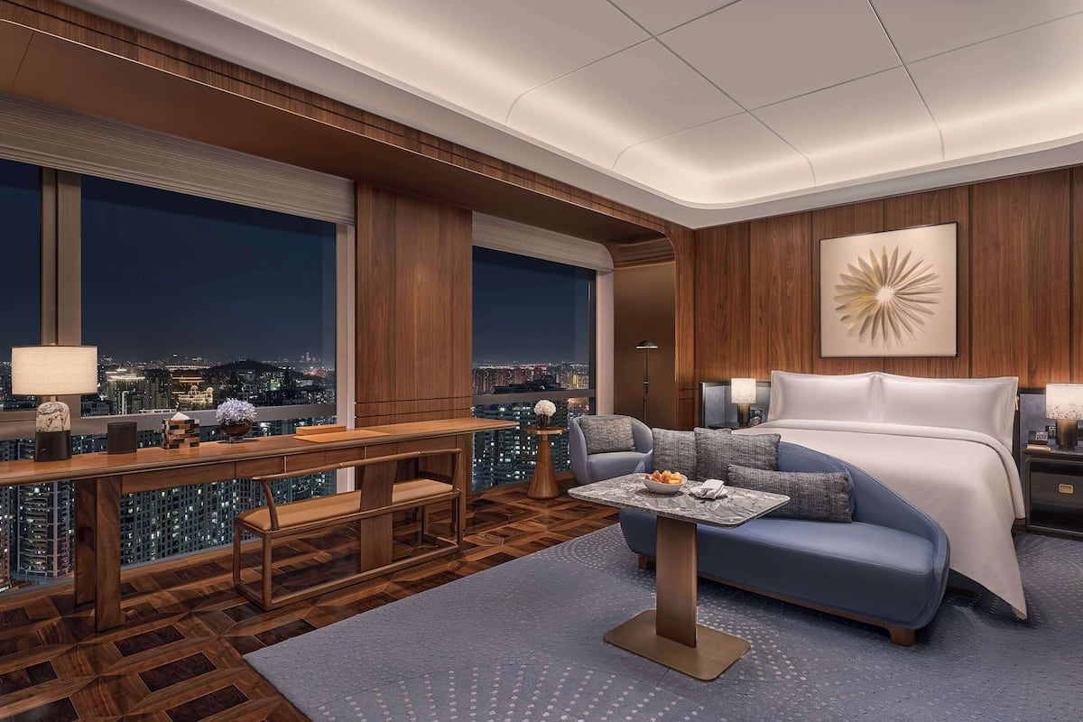 Ein modernes Hotelzimmer mit einem großen Bett, einem Schreibtisch mit Lampe, einer Sitzecke und Blick auf die beleuchtete Stadt bei Nacht.