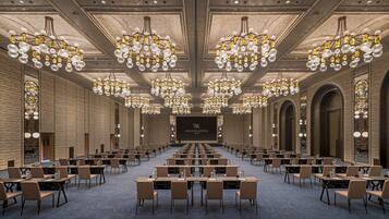 A conference room with rows of tables and chairs, large chandeliers, and a stage with a screen.
