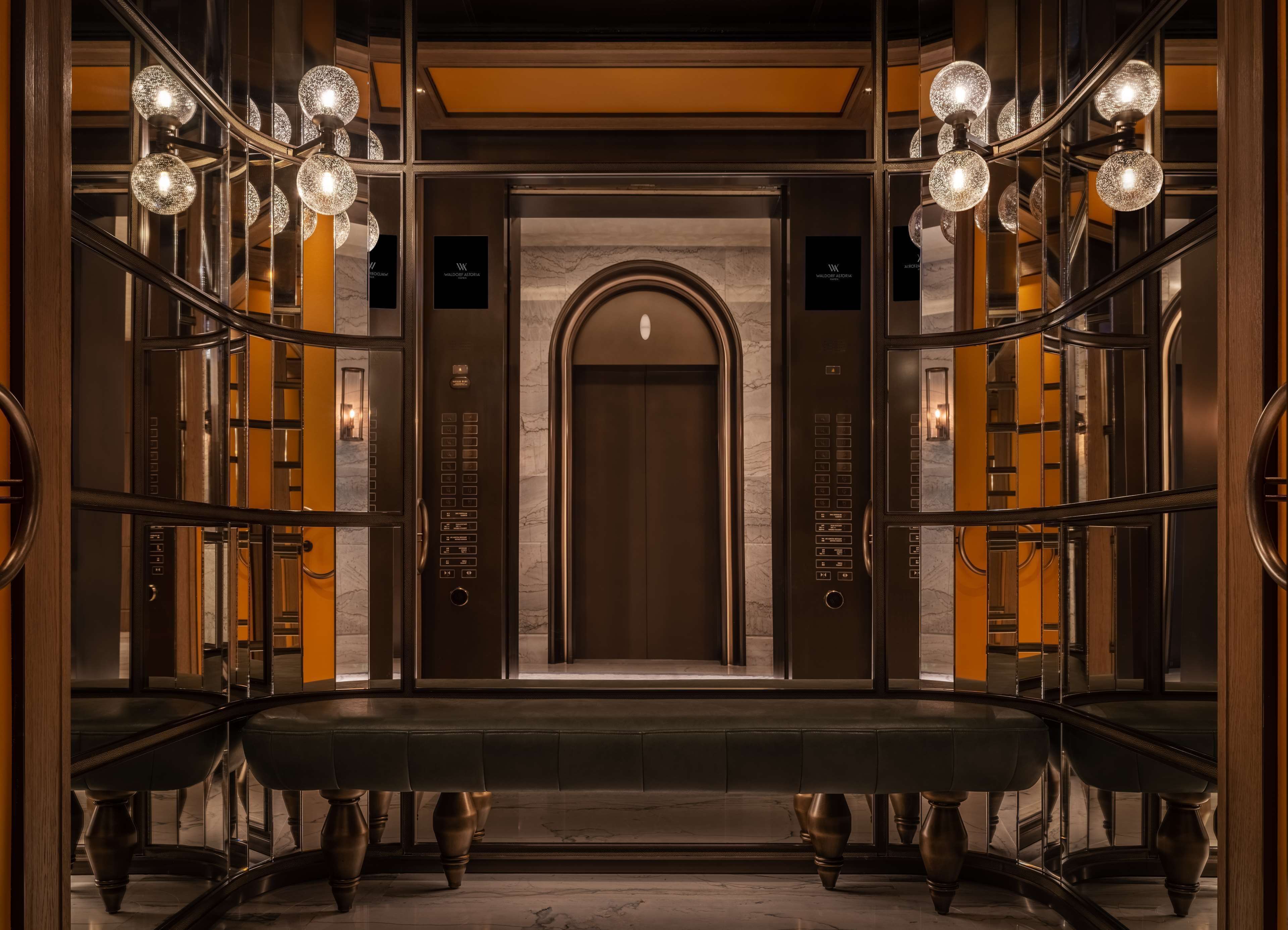 A grand elevator with a marble wall and a central door, flanked by symmetrical lighting fixtures and a bench.