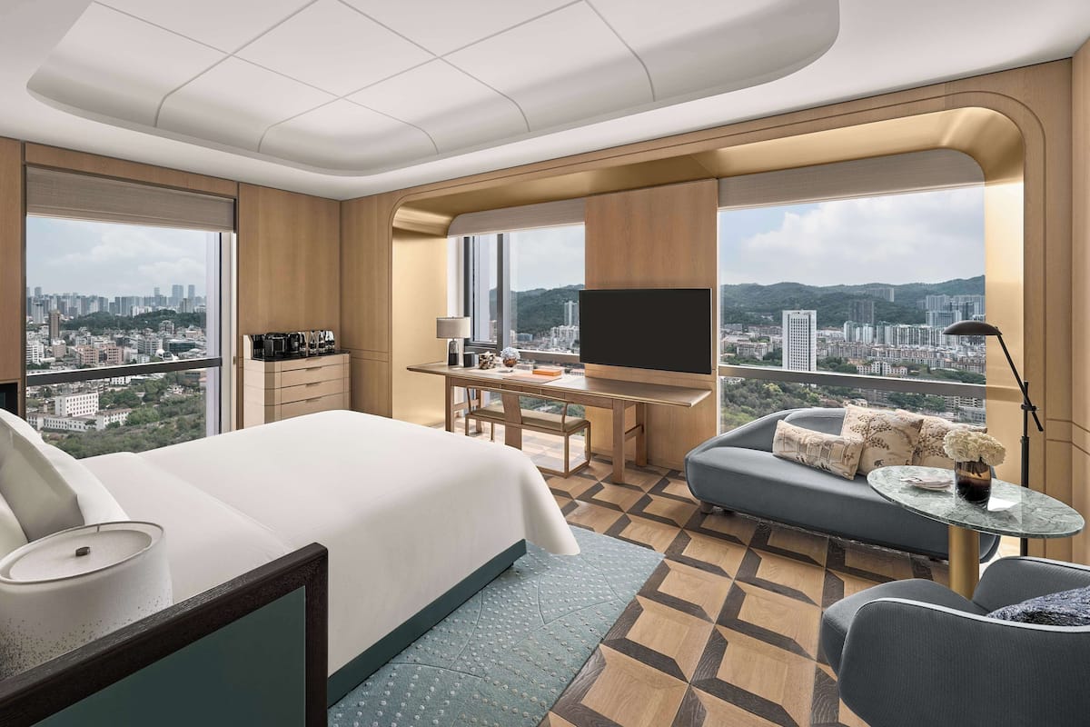 Ein Hotelzimmer mit einem großen Bett, einem Schreibtisch, einer Couch und einem Fernseher. Das Zimmer verfügt über große Fenster mit Blick auf die Stadt.
