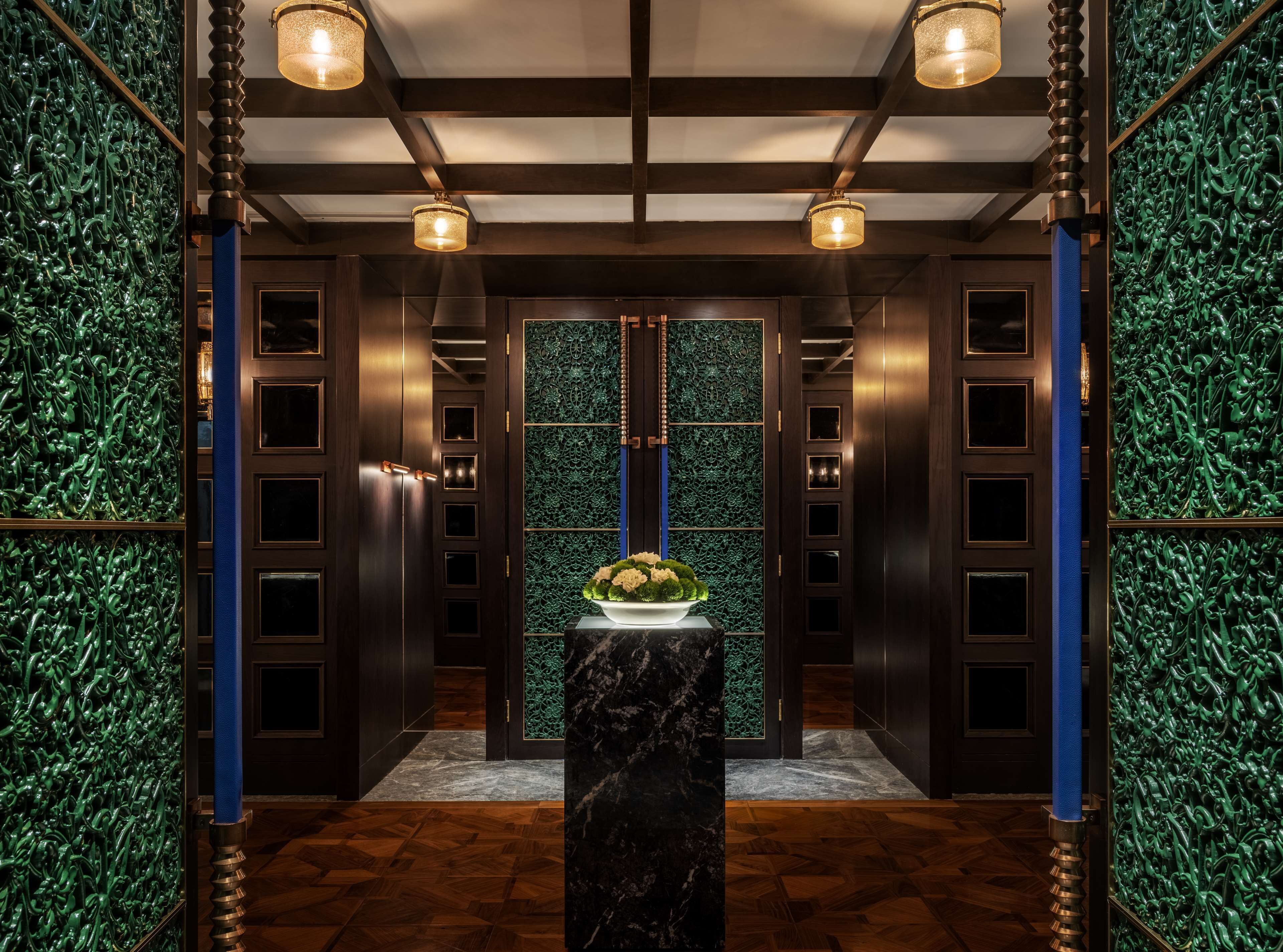 A hallway with dark wooden doors, green patterned walls, and a marble pedestal with a vase of flowers.
