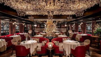 A luxurious dining area with round tables, red upholstered chairs, and a large, ornate chandelier.