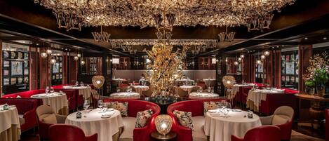 A luxurious dining area with round tables, red upholstered chairs, and a large, ornate chandelier.