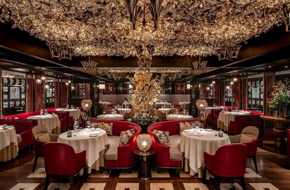 A luxurious dining area with round tables, red upholstered chairs, and a large, ornate chandelier.