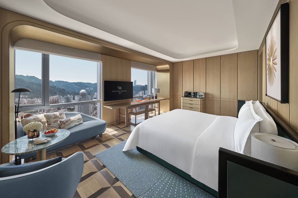 Ein modernes Hotelzimmer mit einem großen Bett, einer Couch, einem Schreibtisch mit Fernseher und Blick auf die Stadt.