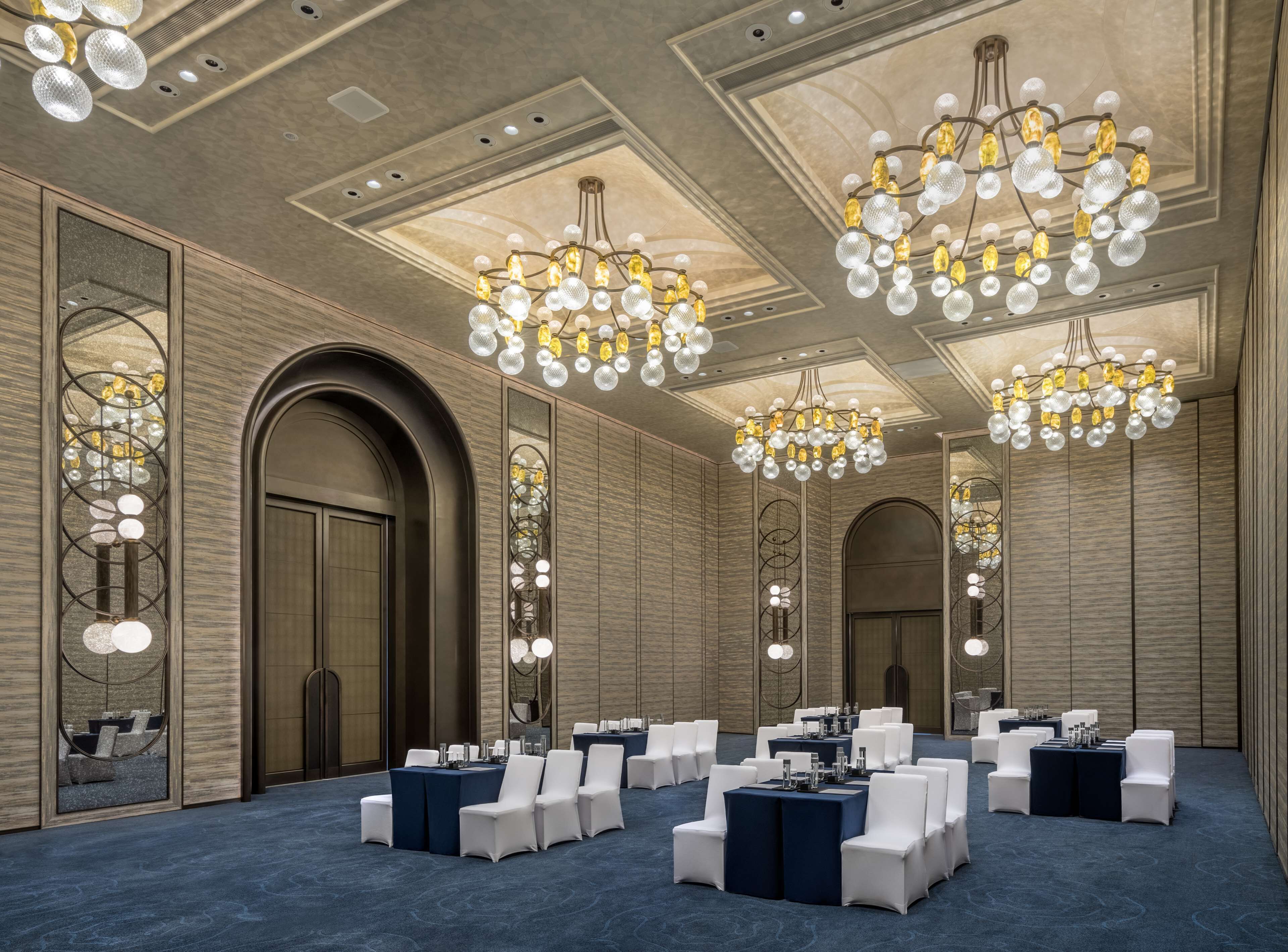 A conference room with multiple tables and chairs, large chandeliers, and a high ceiling with recessed lighting.