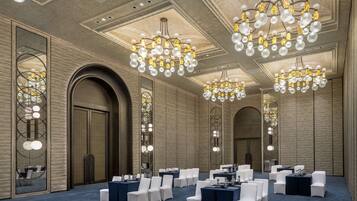 A conference room with multiple tables and chairs, large chandeliers, and a high ceiling with recessed lighting.