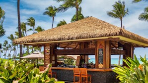 Un bar situé en bord de mer, avec un toit de chaume, des chaises en bois et un tableau affichant le menu.