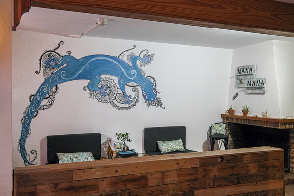 A room with a wooden reception desk, two black sofas with patterned cushions, a decorative wall mural, and a fireplace with potted plants. - Mana Mana Youth Hostel (Tossa de Mar)