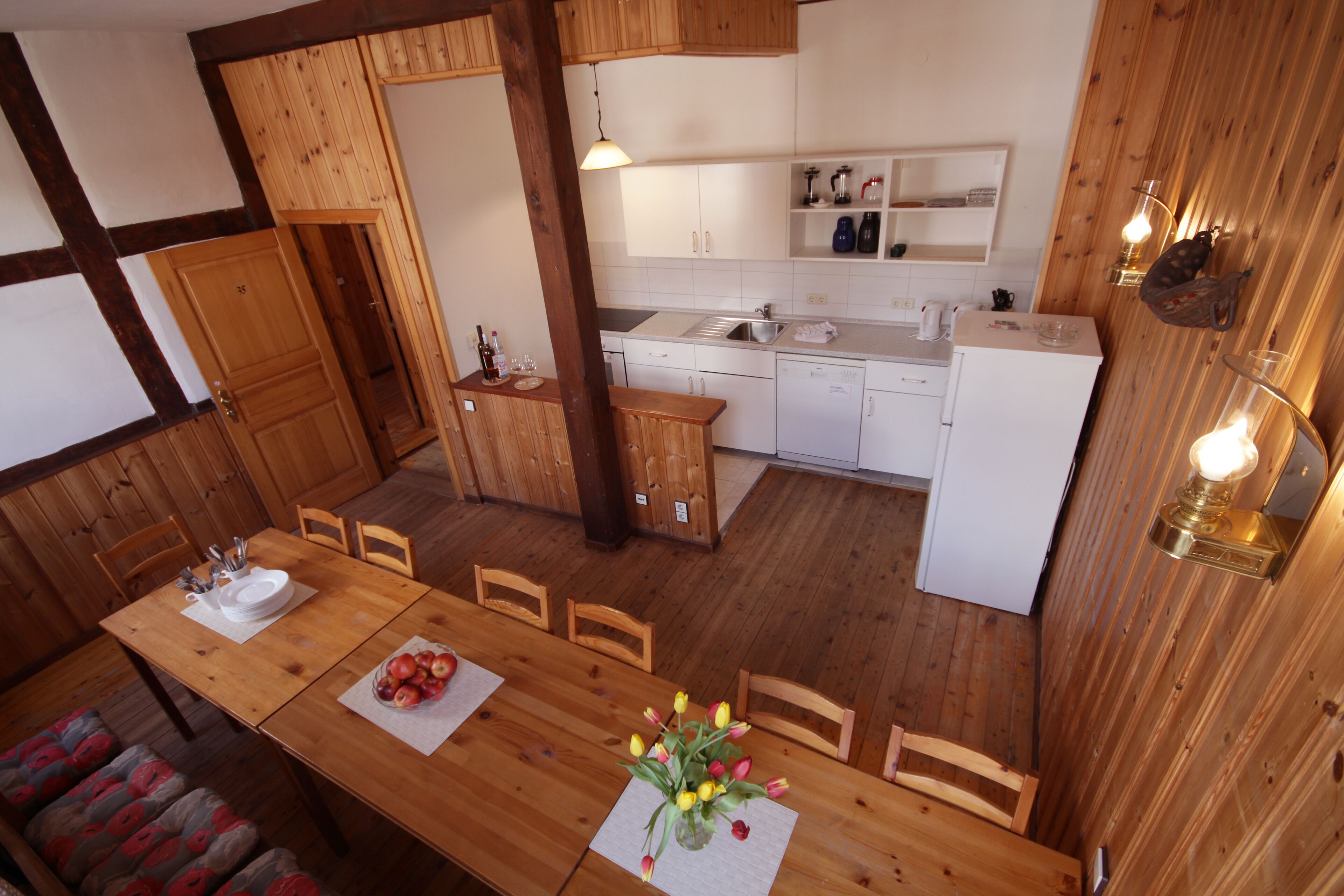 A wooden dining area with a table set for two, a kitchen in the background, and a wooden wall with a light fixture.