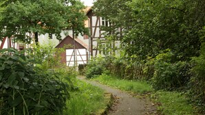 A narrow cobblestone path flanked by dense foliage and a traditional half-timbered building.