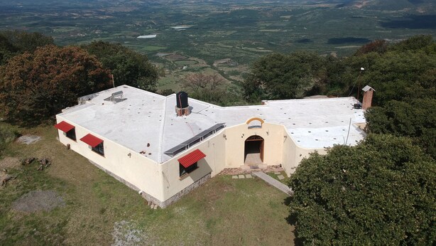 A building with a flat roof and a small entrance. It is surrounded by greenery and has a view of distant hills and fields.